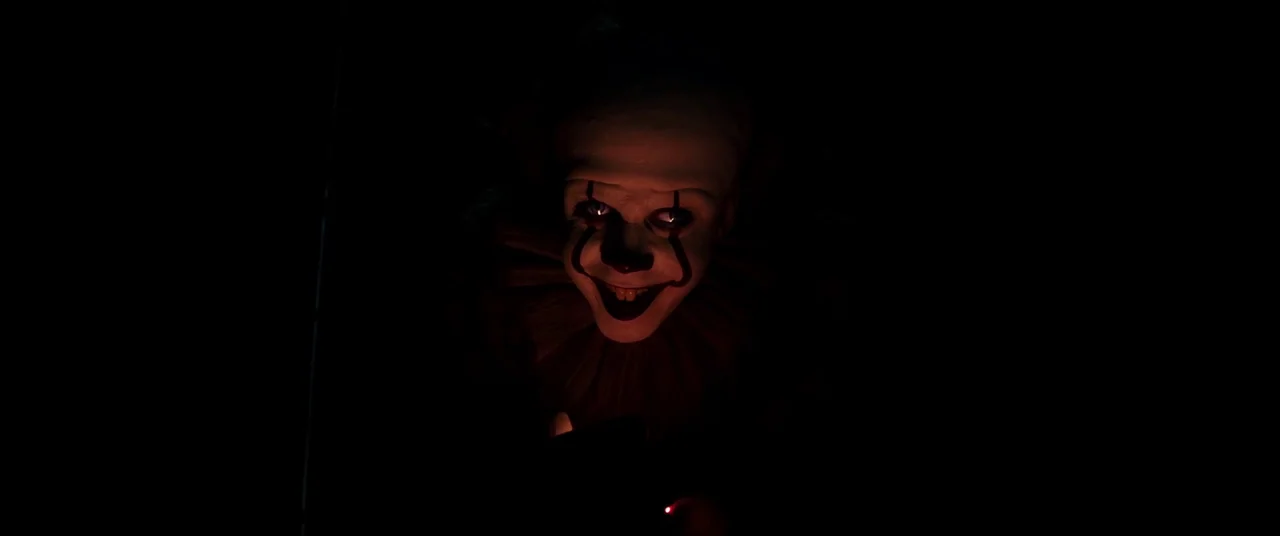 It Chapter Two (2019) movie still: closeup — A character with white face paint and a frilled collar emerges from darkness. The clown stares directly…
