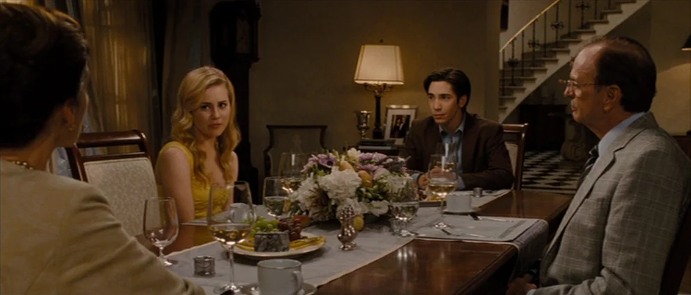 Drag Me to Hell (2009) movie still: wide — A formal dining room features a long wooden table with a large floral centerpiece. Three people are…