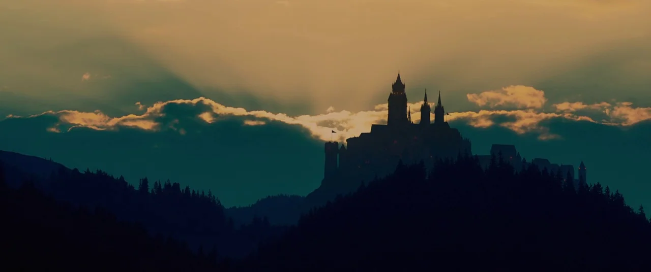 Ophelia (2018) movie still: establishing — A dark, silhouetted castle stands prominently on a mountain ridge against a sunset sky. Dense layers of…