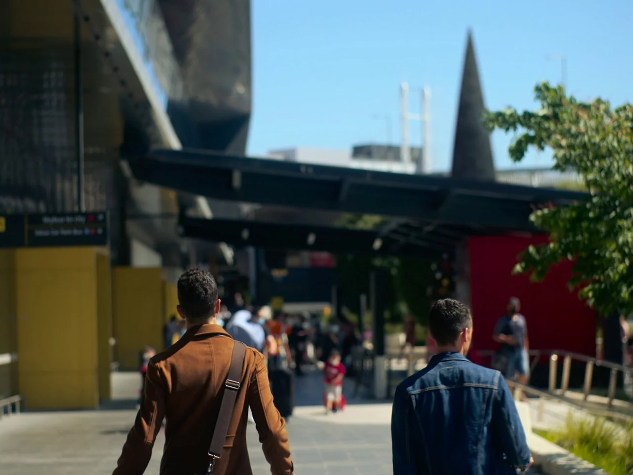 Of An Age (2022) movie still: medium — A modern outdoor pedestrian walkway featuring a tall black spire and red accent walls. Two men walk away from…