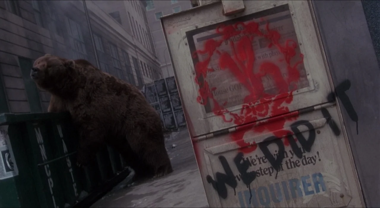 Twelve Monkeys (1995) movie still: wide — A brown bear stands by a green dumpster on a grey urban sidewalk. A metal newspaper rack with red and…