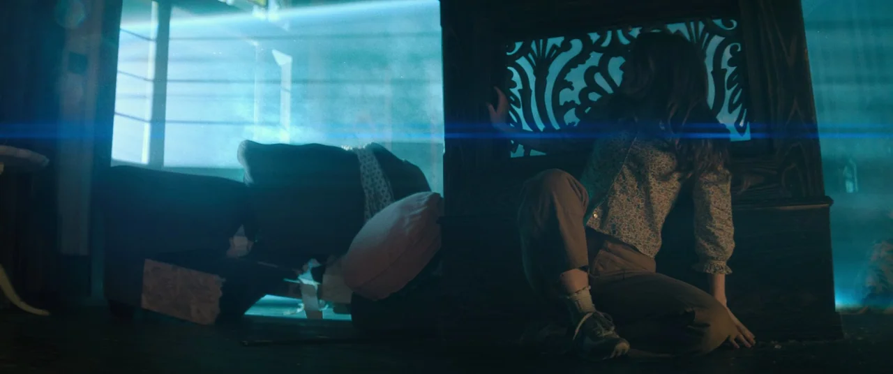No One Will Save You (2023) movie still: medium — A dark space with a wooden decorative partition and an overturned sofa in back. A young woman sits…