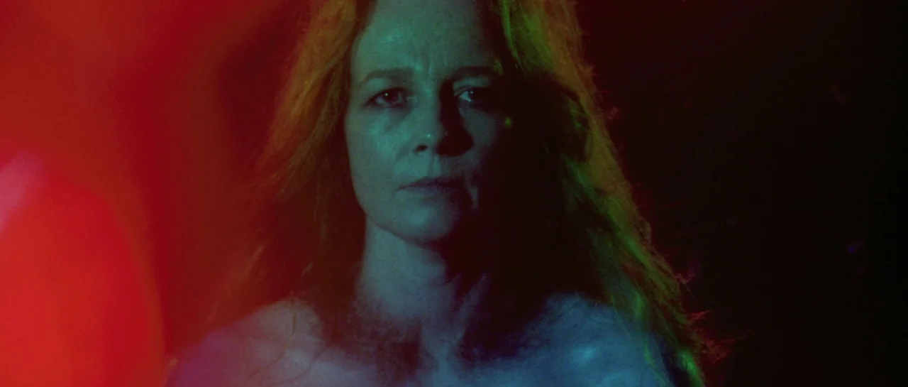 After Blue (2021) movie still: closeup — A woman is framed in extreme close-up within a dimly lit environment. Vivid red and green lights contrast…