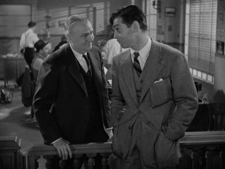 It Happened One Night (1934) movie still: medium — A busy 1940s newsroom features wooden partitions and desk areas with rows of blinds. Two men in…