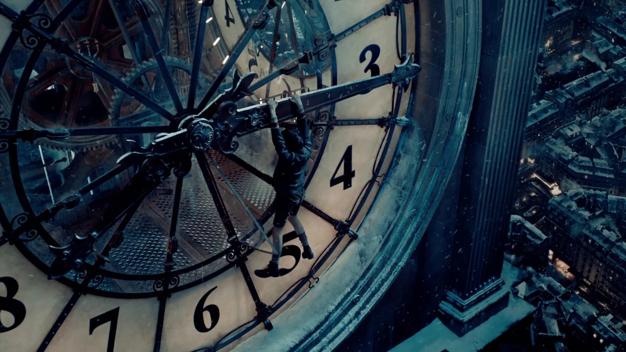 Hugo (2011) movie still: wide — A young boy clings to the large metal hand of an outdoor clock face. Below the mechanism, a dense urban city is…