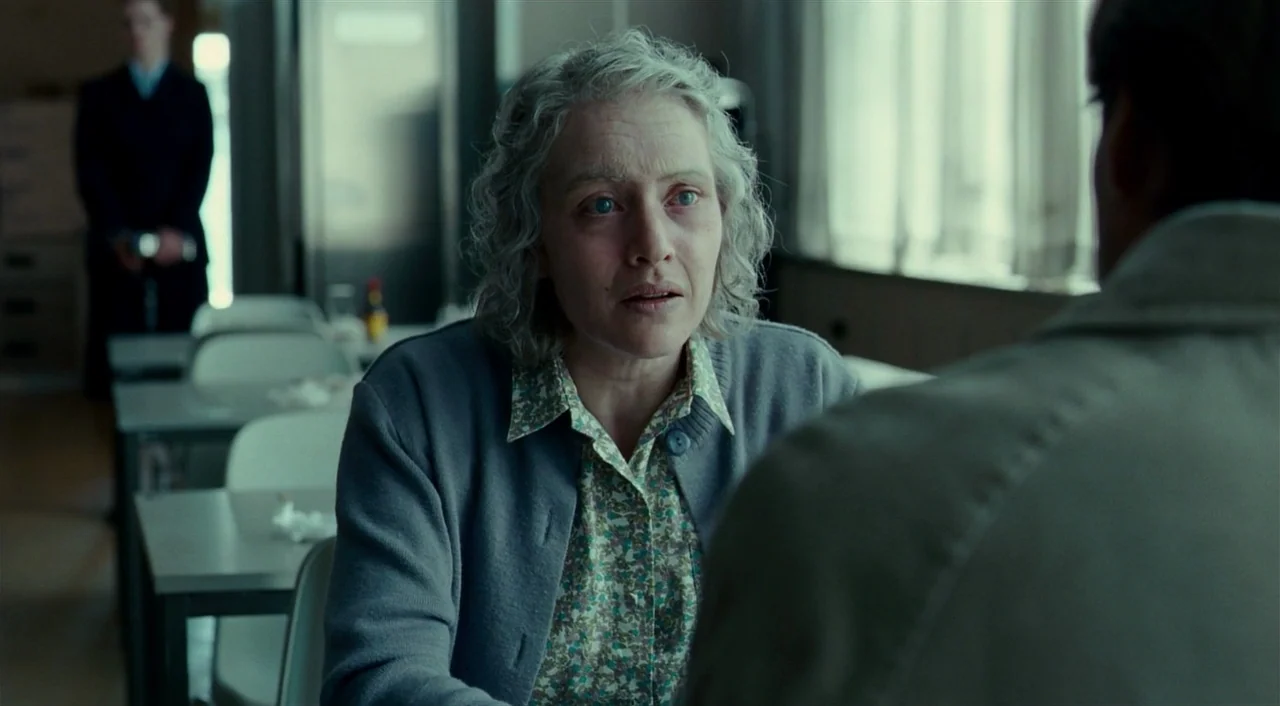 The Reader (2008) movie still: medium — A quiet cafeteria area with rows of metal tables and white chairs. A woman with grey hair sits at a table…