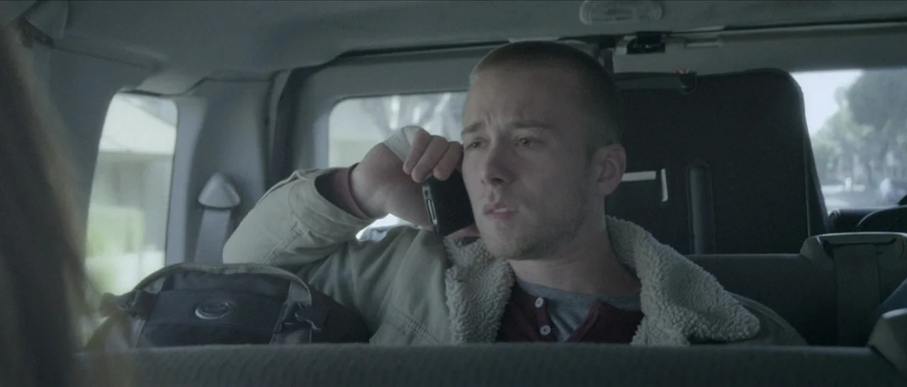 Spring (2014) movie still: medium — The interior of a car shows a man sitting in the back passenger seat. He holds a black smartphone to his ear…