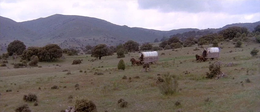 Flesh+Blood (1985) movie still: wide — A wide expanse of hilly rural terrain features scattered shrubs and distant rolling mountains. Two covered…