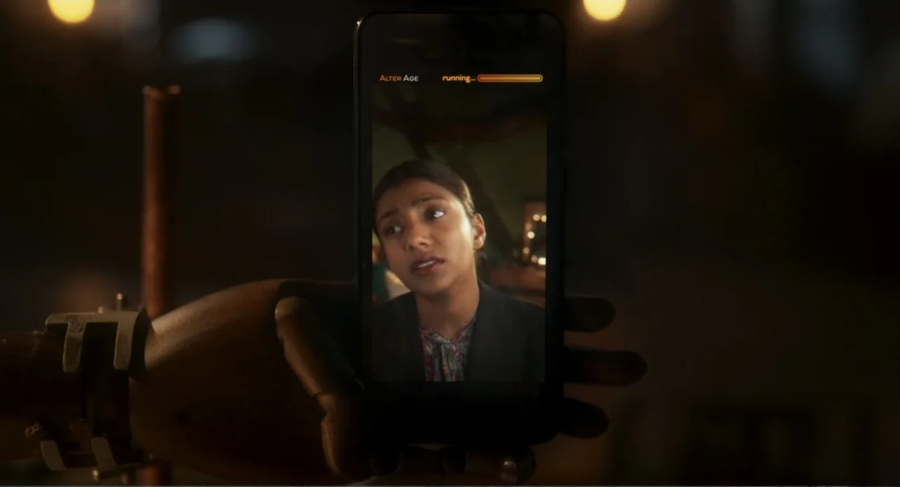 How to Date Billy Walsh (2024) movie still: closeup — A wooden articulated hand holds a smartphone displaying a woman on a screen. The woman's face…