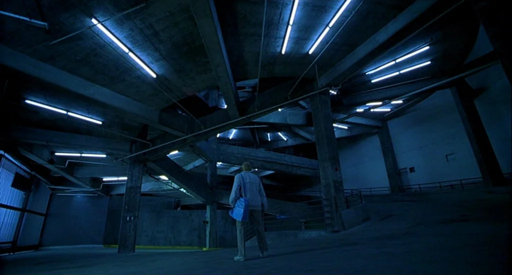 One Hour Photo (2002) movie still: wide — An expansive concrete parking structure features low-hanging ceilings with parallel fluorescent light…