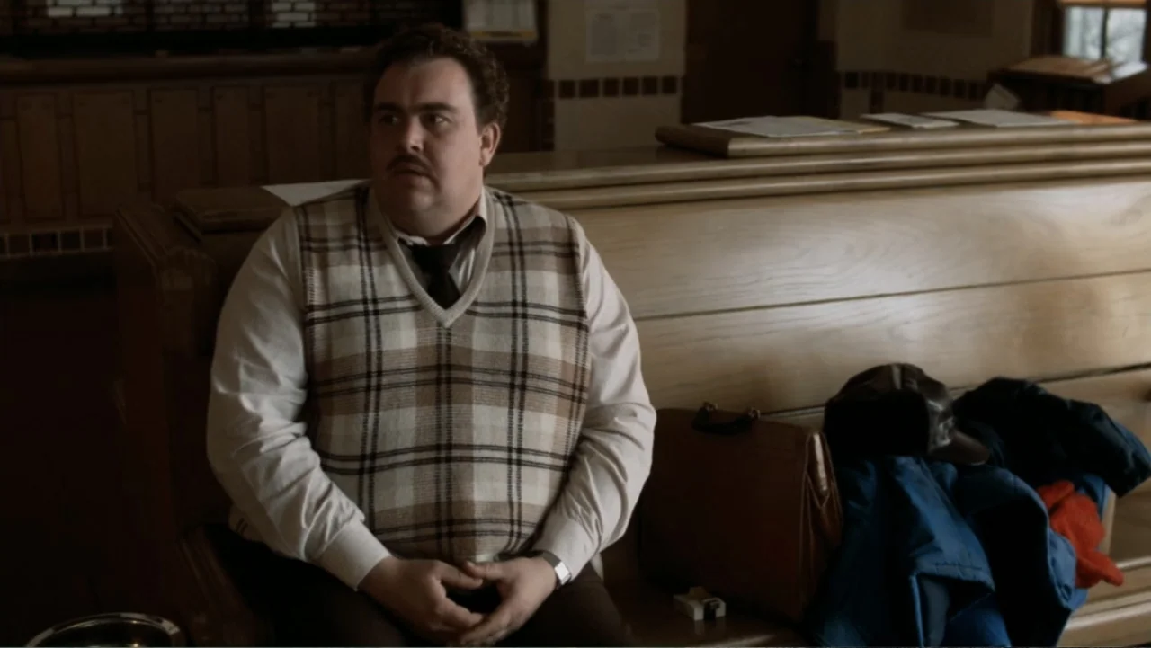 John Candy: I Like Me (2025) movie still: medium — A wooden interior waiting area features a high-backed bench and a leather briefcase. A man in a…