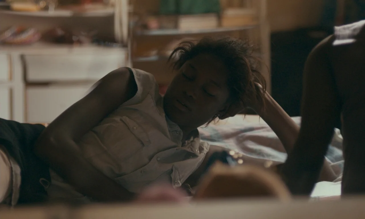 Atlantics (2019) movie still: medium — A cluttered domestic space features a person resting on a bed covered in bedding. The female subject lies…