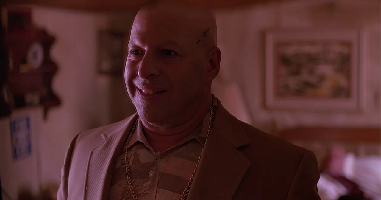 Darkman (1990) movie still: medium — A dimly lit room with a framed picture on the wall behind the subject. A bald man smiles while wearing a tan…