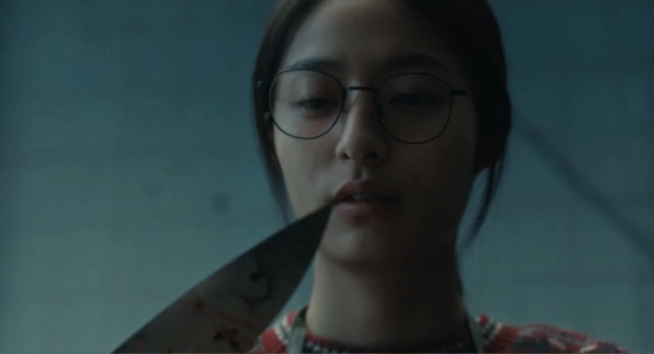 Winter in Sokcho (2025) movie still: closeup — An interior space with a plain wall as the background. A young woman in a patterned sweater holds a…