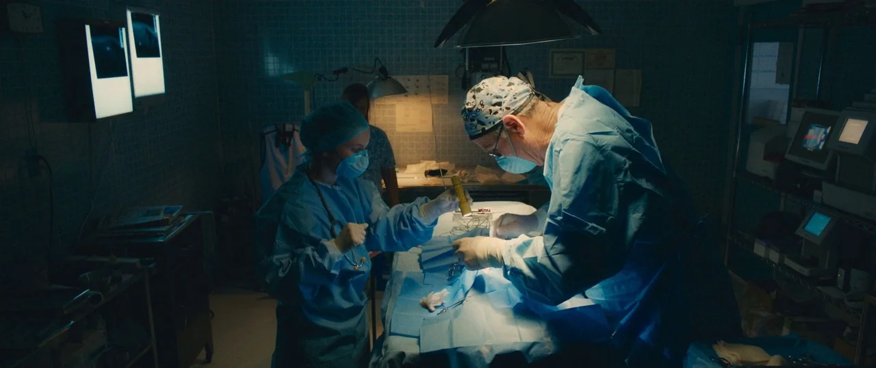 Manglehorn (2014) movie still: medium — A dimly lit surgical room with blue-tiled walls and various medical equipment in the background. A surgeon…