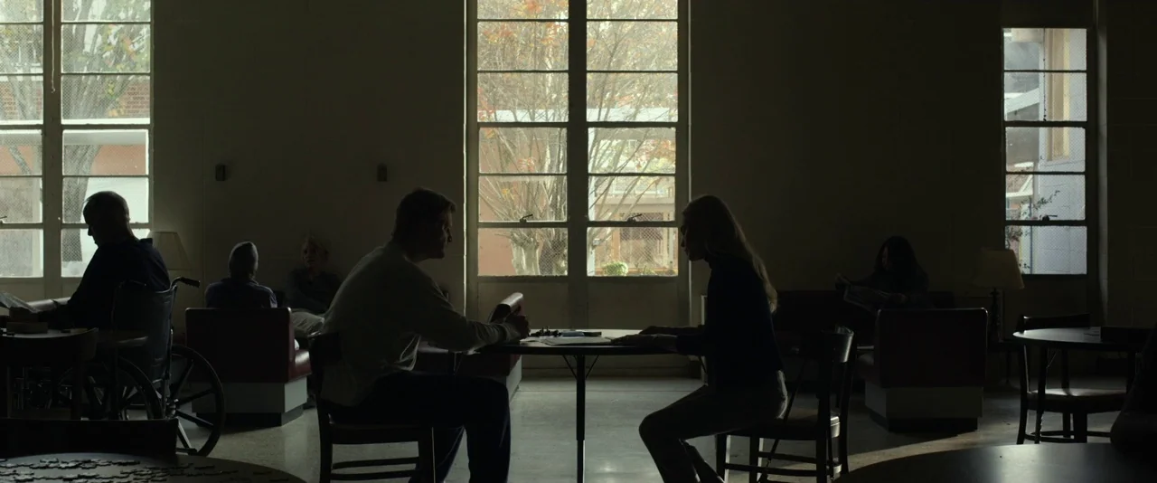 Before I Wake (2016) movie still: wide — A spacious, dimly lit facility common room with large windows and multiple tables. Several people sit…