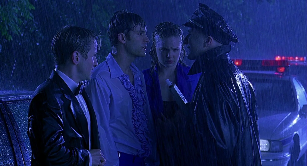 Bride of Chucky (1998) movie still: medium — A group of people are gathered on a wet road at night in the rain. A police officer in a dark coat…