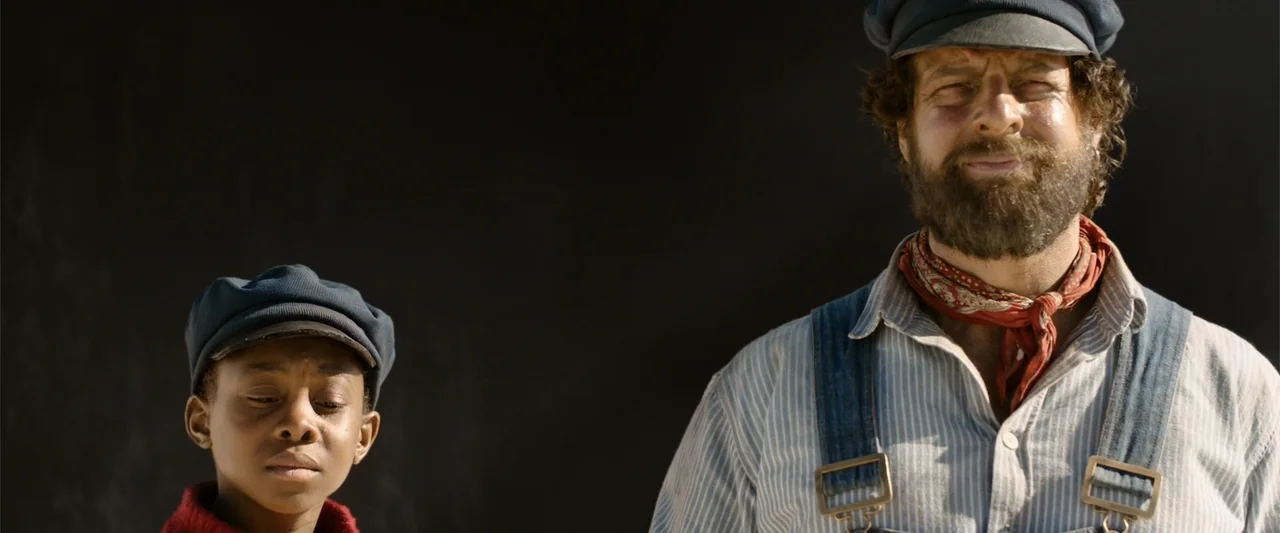 Jim Button and Luke the Engine Driver (2018) movie still: medium — An adult man and a young boy stand before a dark, featureless backdrop. The man…