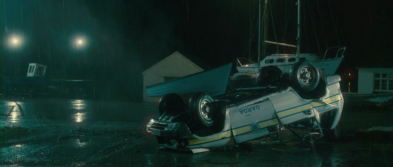 Grabbers (2012) movie still: wide — A rain-drenched dock area at night features a flipped Irish police patrol car. Several small boats and a white…