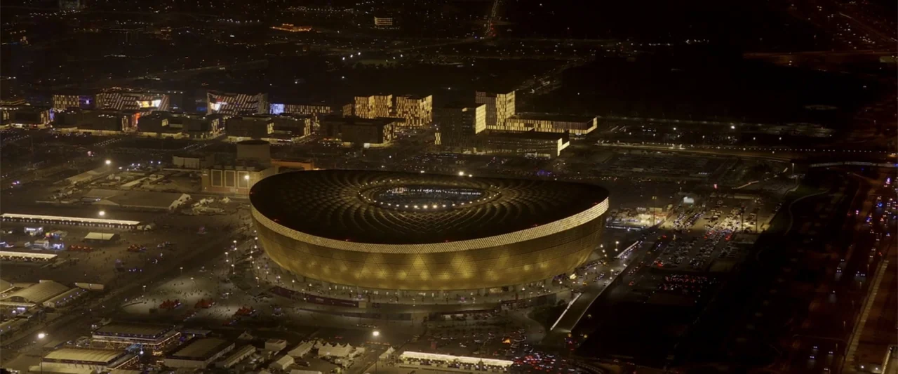 I Choose to Believe (2023) movie still: establishing — An aerial night view shows a large modern stadium with a golden geometric facade. The…