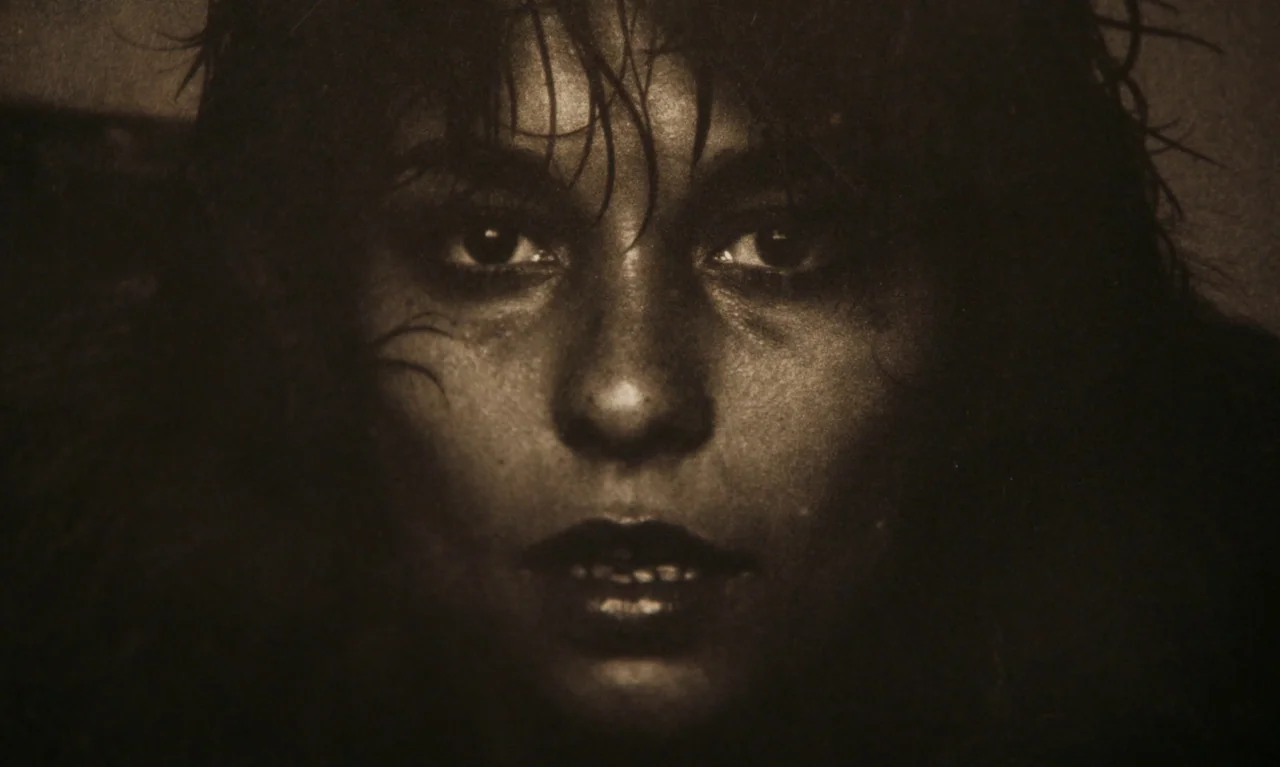 Little Girl Blue (2023) movie still: closeup — A dark, undefined space surrounds a woman's face. The subject stares forward with wet hair framing…