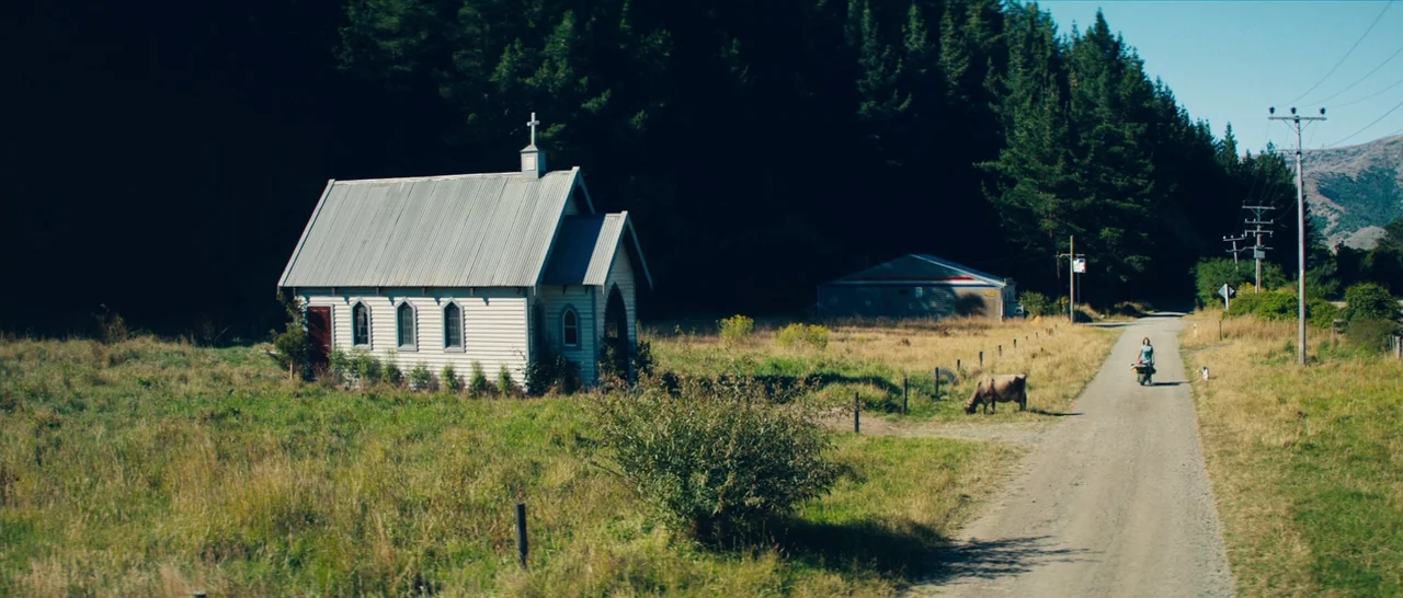 Z for Zachariah (2015) movie still: wide — A small white wooden chapel sits in a grassy field beside a dirt road. A person walks down the path…