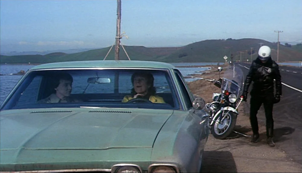 Harold and Maude (1971) movie still: medium — A light green sedan sits on the shoulder of a road bordering a coastal area. A police officer in…
