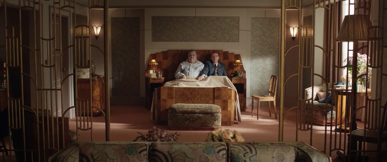 Domino (2005) movie still: wide — A spacious, symmetrical hotel suite decorated in an Art Deco style with ornate metal screens. Two men sit in the…