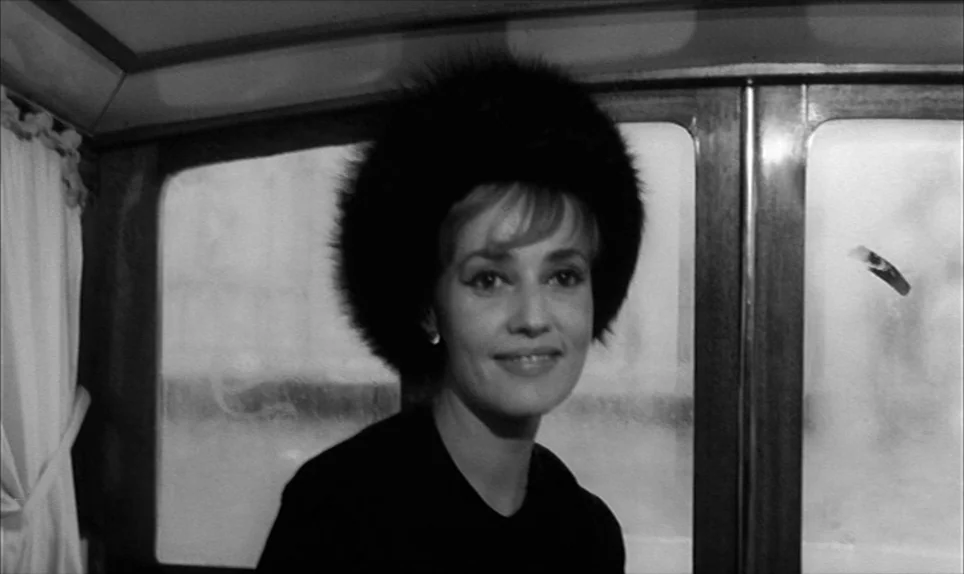 Eva (1962) movie still: medium — A train cabin interior features wooden paneling and windows showing blurred white light. A woman wearing a fur hat…