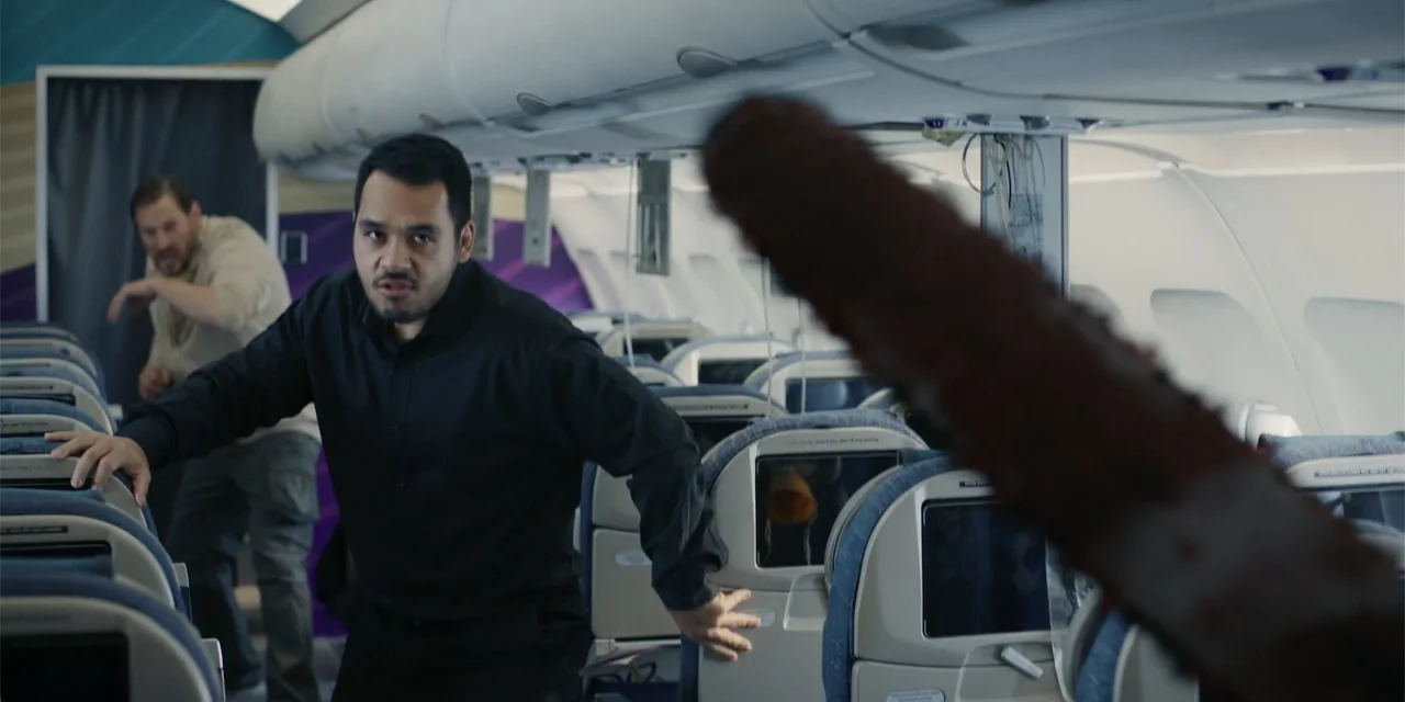 Fight or Flight (2025) movie still: medium — The interior of an airplane cabin features rows of blue and beige passenger seats. Two men stand in the…