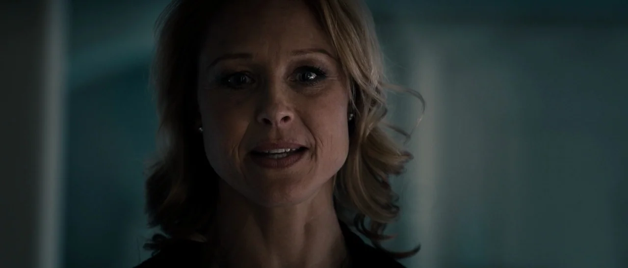 The Purge (2013) movie still: closeup — A dimly lit indoor space with indistinct backgrounds in deep shadow. A woman with blonde hair gazes forward…