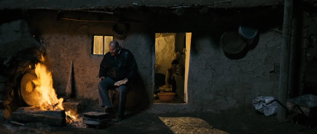 Once Upon a Time in Anatolia (2011) movie still: wide — A dark rural exterior features a mud-walled building at night with a fire burning outside. A…