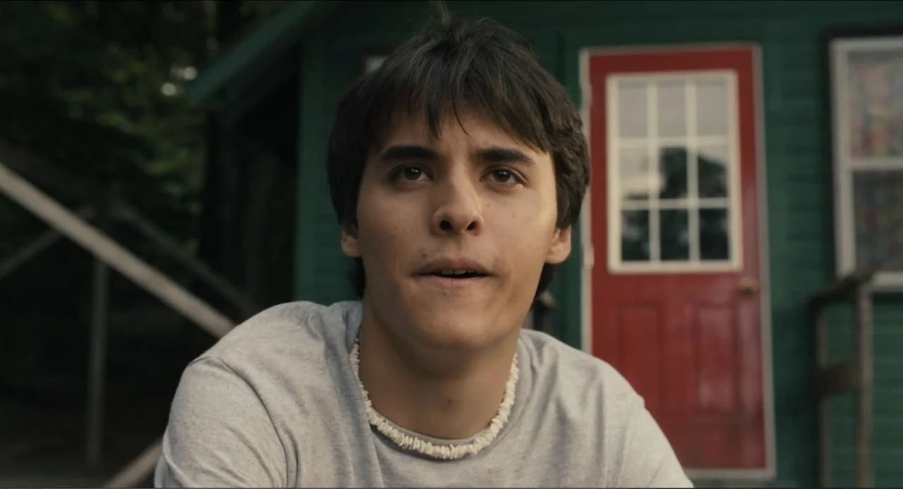 Hell of a Summer (2025) movie still: medium — A dark green wooden building with a prominent red door serves as the backdrop. A young man wearing a…