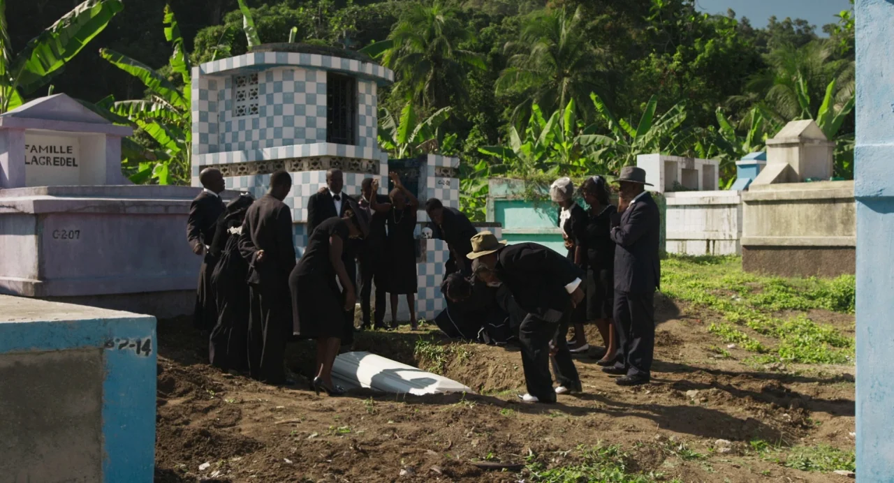 Zombi Child (2019) movie still: wide — A group of mourners in black formal wear gathers at an outdoor burial site. Concrete tombs with checkerboard…
