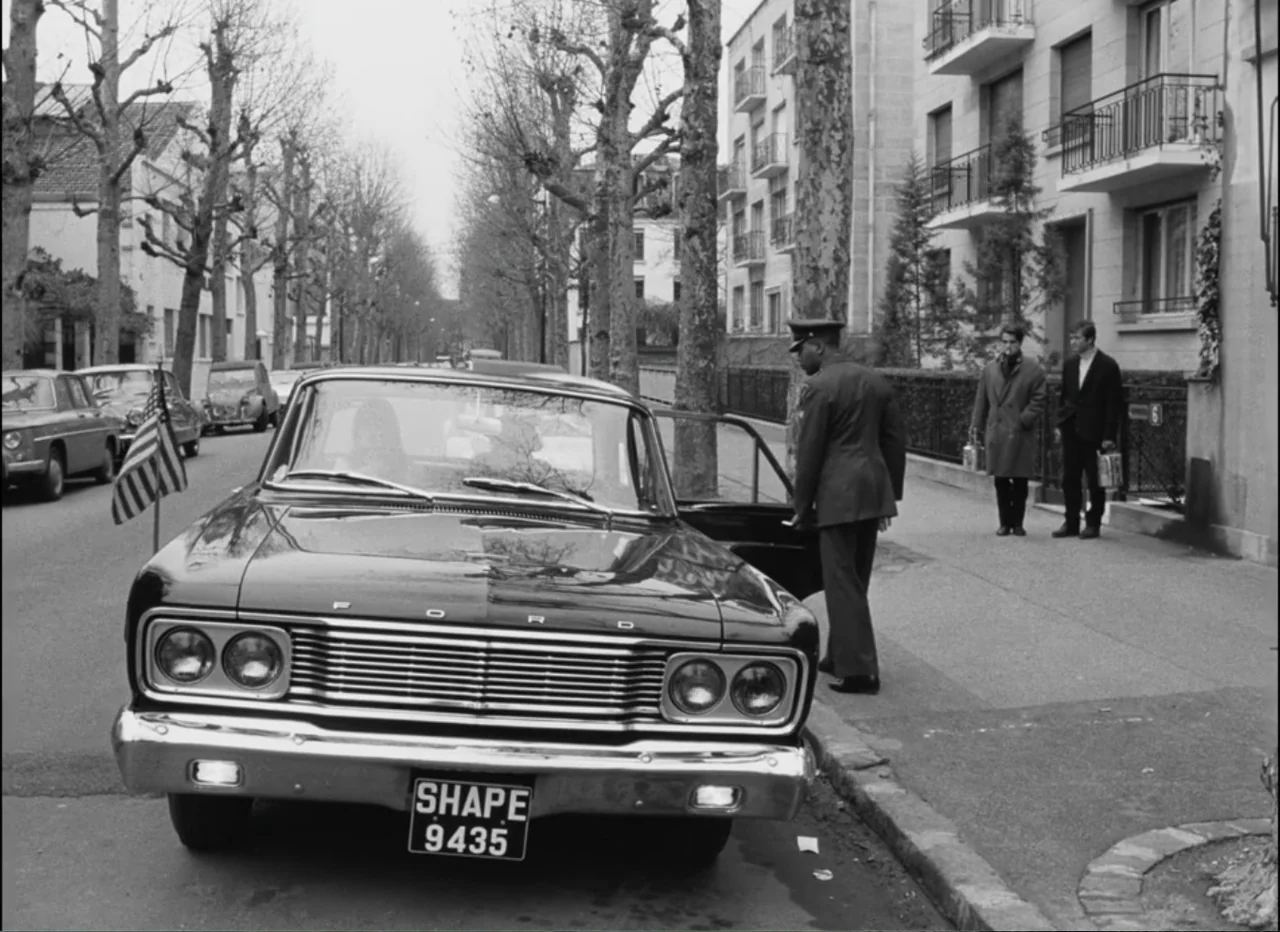 Masculin Féminin (1966) movie still: medium — A quiet residential street features a parked vintage Ford sedan beside a sidewalk. A uniformed man…