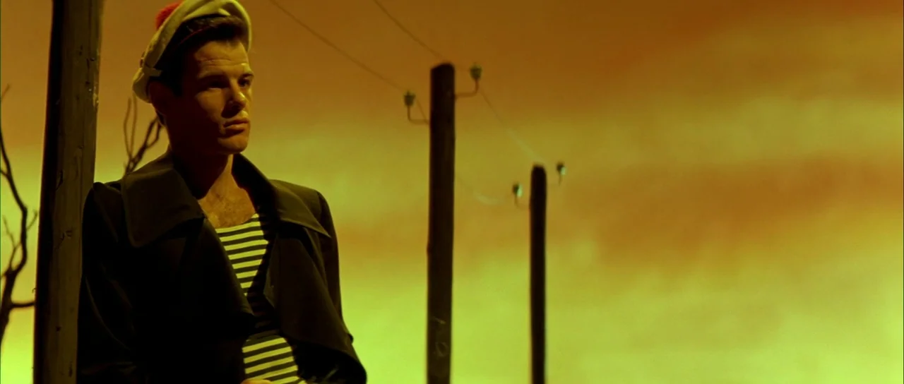 Querelle (1982) movie still: medium — A man in a nautical outfit stands near a roadside utility pole. An vibrant, amber-toned sky serves as the…