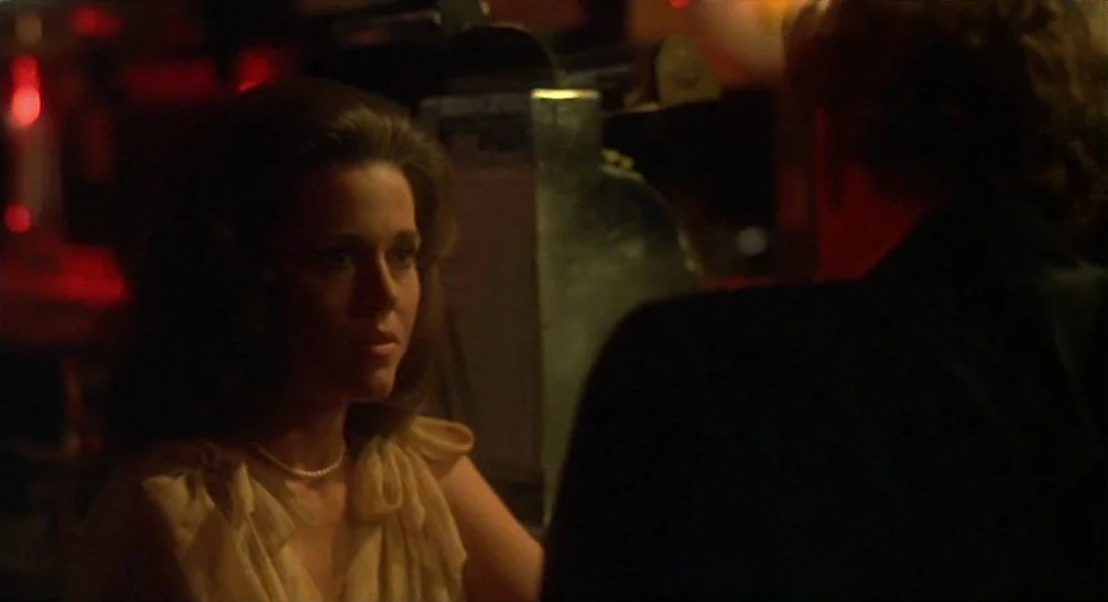 Coming Home (1978) movie still: medium — A dark, dimly lit bar environment with indistinct background shapes and faint red light. A woman in a light…