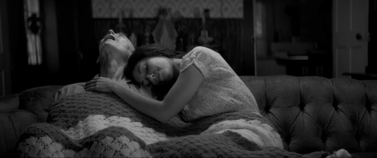 The Eyes of My Mother (2016) movie still: medium — A dark, dimly lit living room with a vintage tufted leather sofa. A woman lies resting her head…