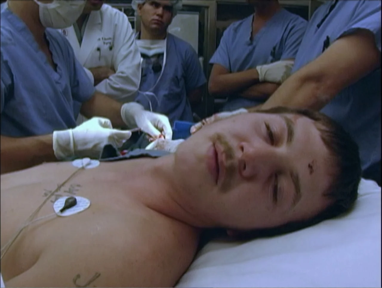 Addiction (2024) movie still: closeup — A sterile hospital operating room filled with medical personnel in scrubs and surgical caps. A young man…