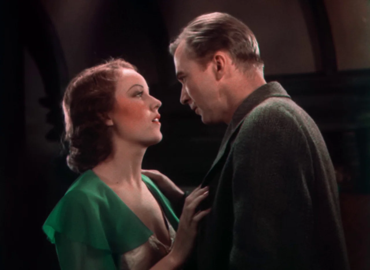 Doctor X (1932) movie still: medium — A dimly lit indoor space with dark, indistinct architectural shadows in the background. A woman in a green…