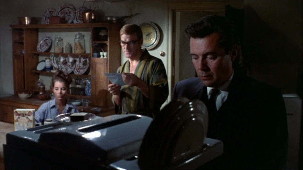Accident (1967) movie still: medium — A rustic kitchen features dark wood furniture and shelves stocked with various dishes. One man in a suit reads…