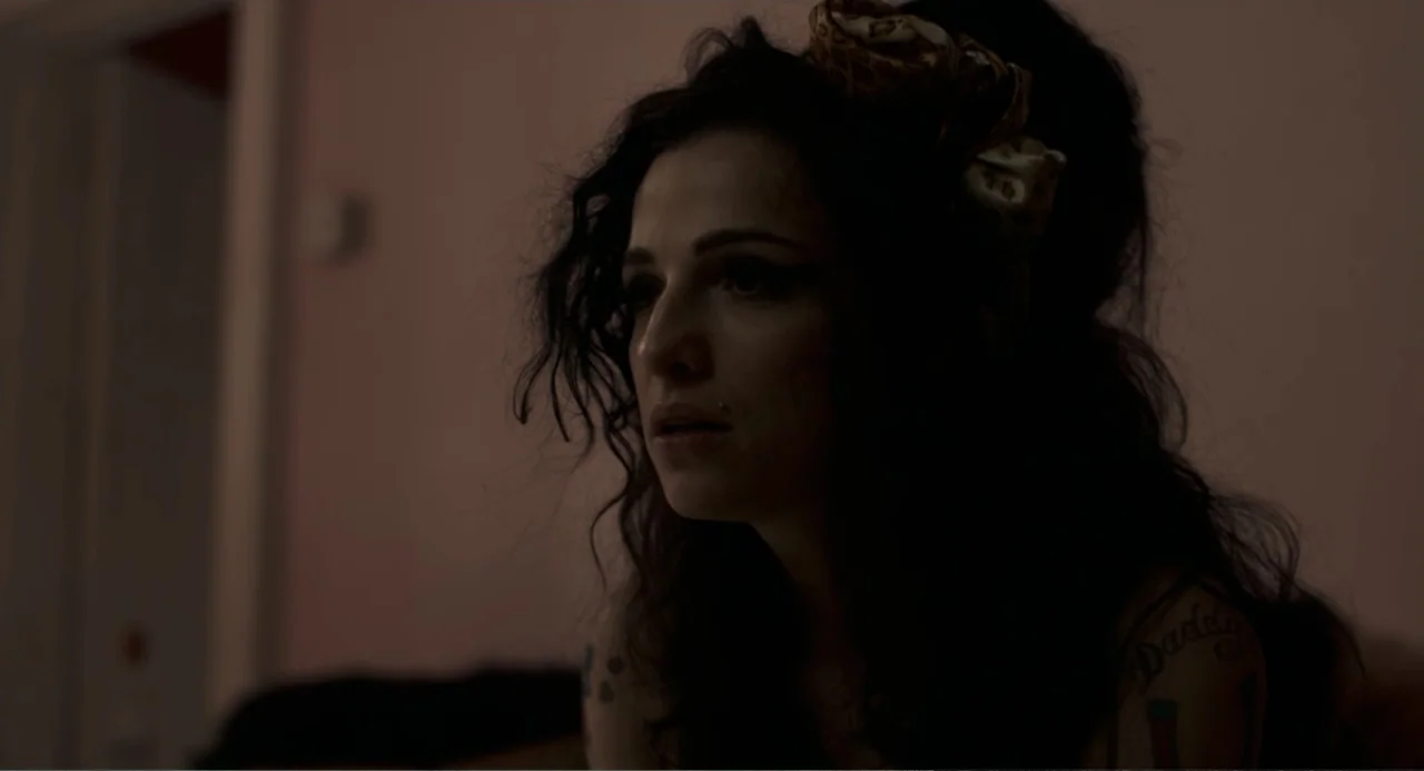 Back to Black (2024) movie still: closeup — A dimly lit room with muted pink walls and low light conditions. A woman with a beehive hairstyle and…