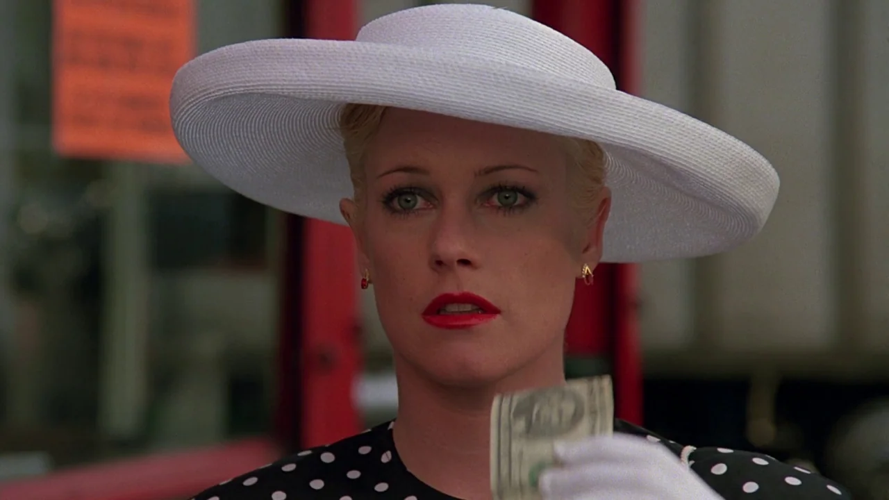 Something Wild (1986) movie still: closeup — A woman in a white hat stands outdoors in front of a blurred urban background. She holds up a single…