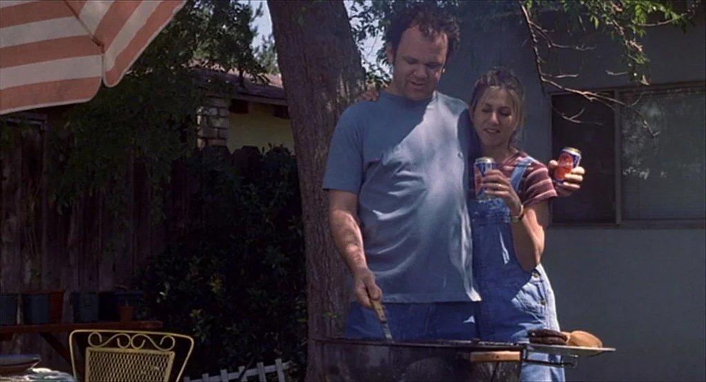 The Good Girl (2002) movie still: medium — A residential backyard features a wooden fence, a tree, and a grill setup. A man in a blue t-shirt grills…