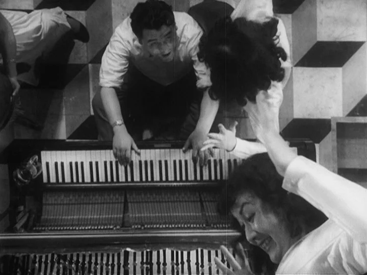 Ikiru (1952) movie still: medium — A top-down view shows a room with a distinct cubic geometric tile floor pattern. A man plays an upright piano…