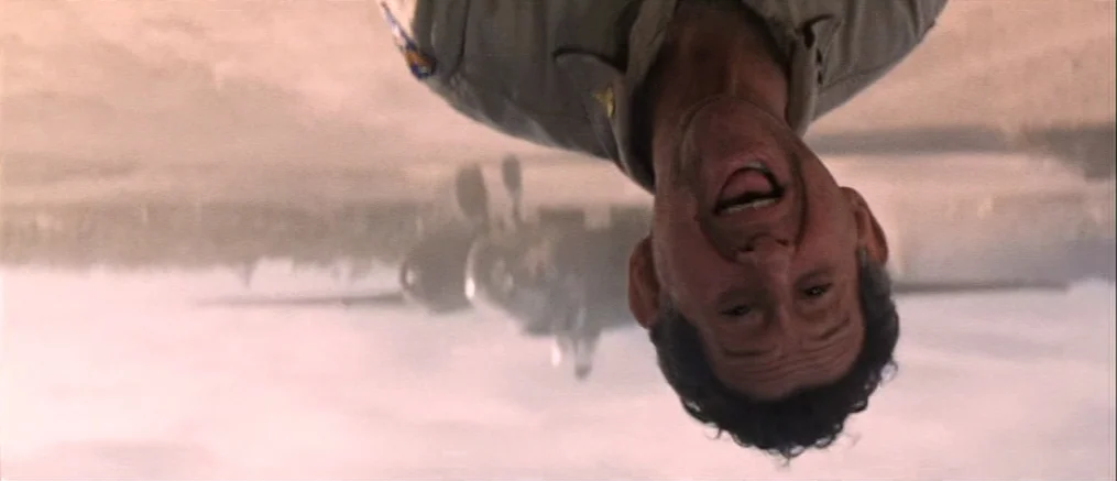 Catch 22 (1970) movie still: closeup — An upside-down shot captures a man laughing on a sunlit military airfield. An airplane sits in the…