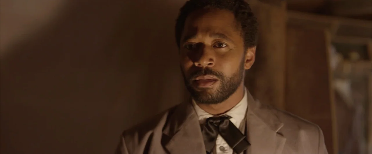 Ghosts of the Ozarks (2022) movie still: closeup — A man stands inside a room with dark, indistinct walls and warm ambient light. He wears a formal…