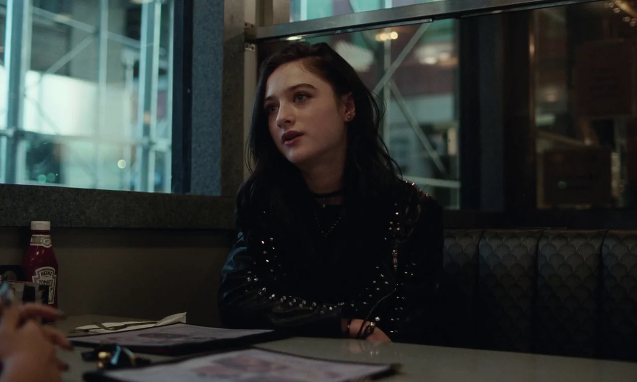 Vox Lux (2018) movie still: medium — A dimly lit diner booth at night with a large window in the background. A young woman with dark hair sits…