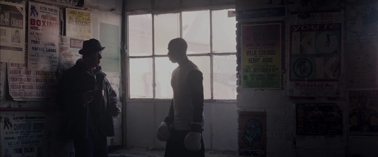Creed (2015) movie still: medium — A dimly lit room with walls covered in vintage boxing posters and flyers. A man in a hat and a boxer in gloves…