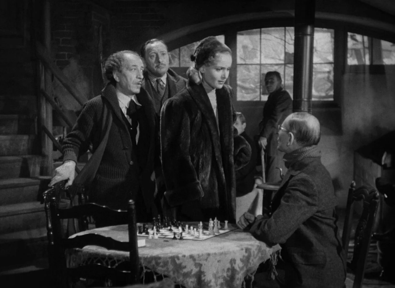 To Be or Not To Be (1942) movie still: medium — A dark tavern room features a wooden table where people gather to watch chess. A woman in a fur coat…
